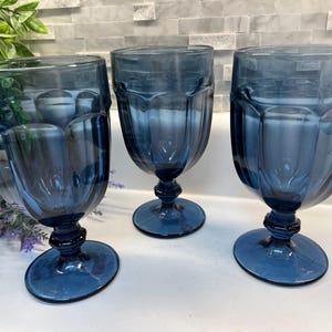 May include: Three matching goblets crafted from translucent blue glass. Each piece features a rounded bowl with vertical paneling, a short decorative stem with a central knob, and a wide circular base for stability. The glass has a deep blue hue.