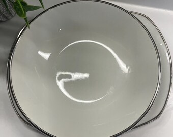 Puede incluir: Un tazón de cerámica blanco con un borde plateado. El tazón está vacío y tiene una superficie reflectante.