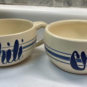 May include: Two off-white ceramic chili bowls with blue stripes and the word "Chili" in a script font. Each bowl has a handle and a rounded shape, perfect for serving soup or stew.