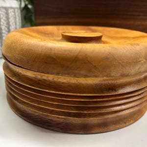 May include: A round, wooden container with a lid. The container is made of light brown wood and has a ridged, textured design on the sides. The lid has a small, circular handle. The container is likely used for storage.