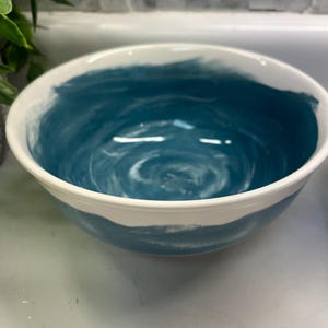 May include: A ceramic bowl with a white rim and a teal blue interior. The bowl's interior features a swirling pattern of white and blue paint. The exterior is white with a band of teal blue paint.