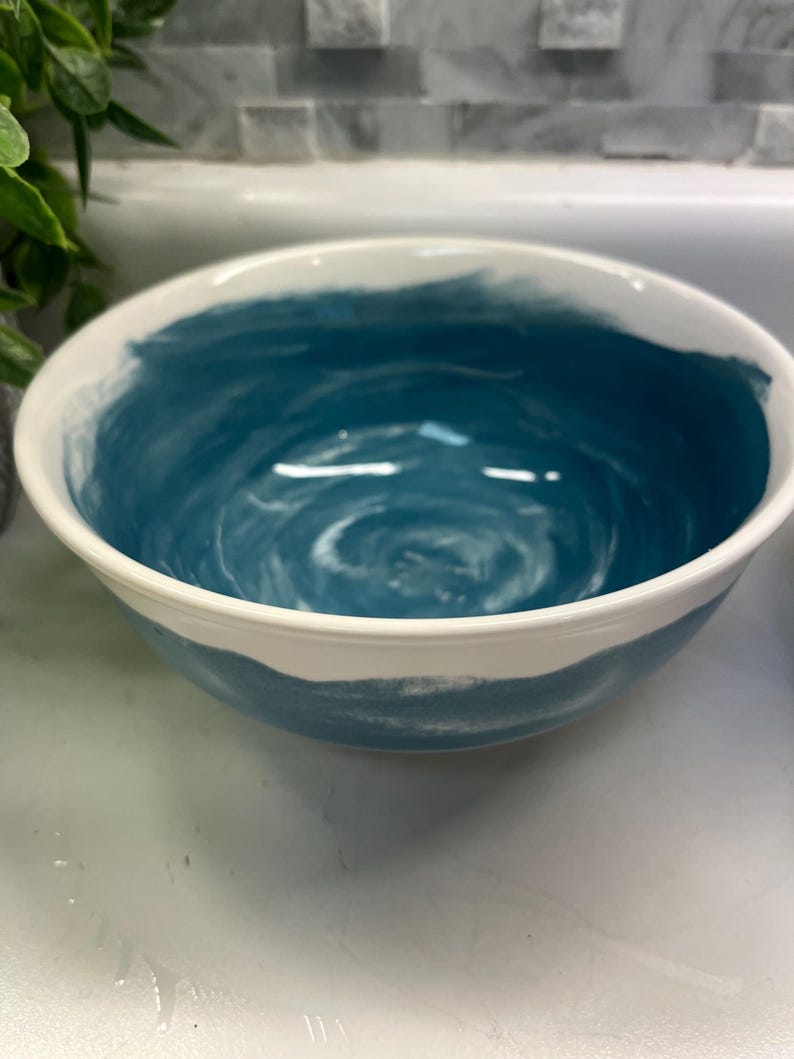 May include: A ceramic bowl with a white rim and a teal blue interior. The bowl's interior features a swirling pattern of white and blue paint. The exterior is white with a band of teal blue paint.