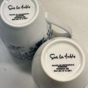 Puede incluir: Dos tazas de cer&aacute;mica blanca con dise&ntilde;os decorativos azules. La parte inferior de cada taza presenta el texto "Sur la table", "Made in Indonesia" y "Dishwasher, Microwave and Oven Safe up to 220&deg;C."
