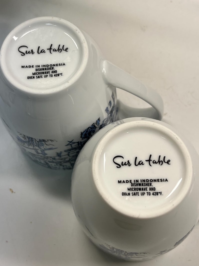 Puede incluir: Dos tazas de cer&aacute;mica blanca con dise&ntilde;os decorativos azules. La parte inferior de cada taza presenta el texto "Sur la table", "Made in Indonesia" y "Dishwasher, Microwave and Oven Safe up to 220&deg;C."