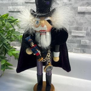 May include: A decorative wooden nutcracker figurine in a top hat and cape. The nutcracker has a white beard and holds a miniature soldier. It has a gold base and a pocket watch charm. The hat is black with a feather and purple accent.