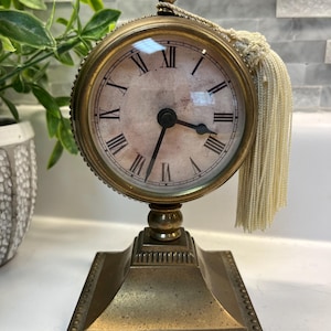 May include: A brass-toned table clock with a circular face featuring black Roman numerals and a weathered, off-white background. The clock is mounted on a square, tiered base and topped with a decorative finial. A long, cream-colored tassel hangs from the top right side of the clock frame.