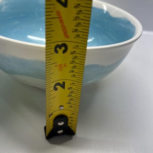 May include: A white ceramic bowl with a light blue interior and a blue painted band around the exterior. The bowl is approximately 7.6 cm deep, as measured by a yellow tape measure.