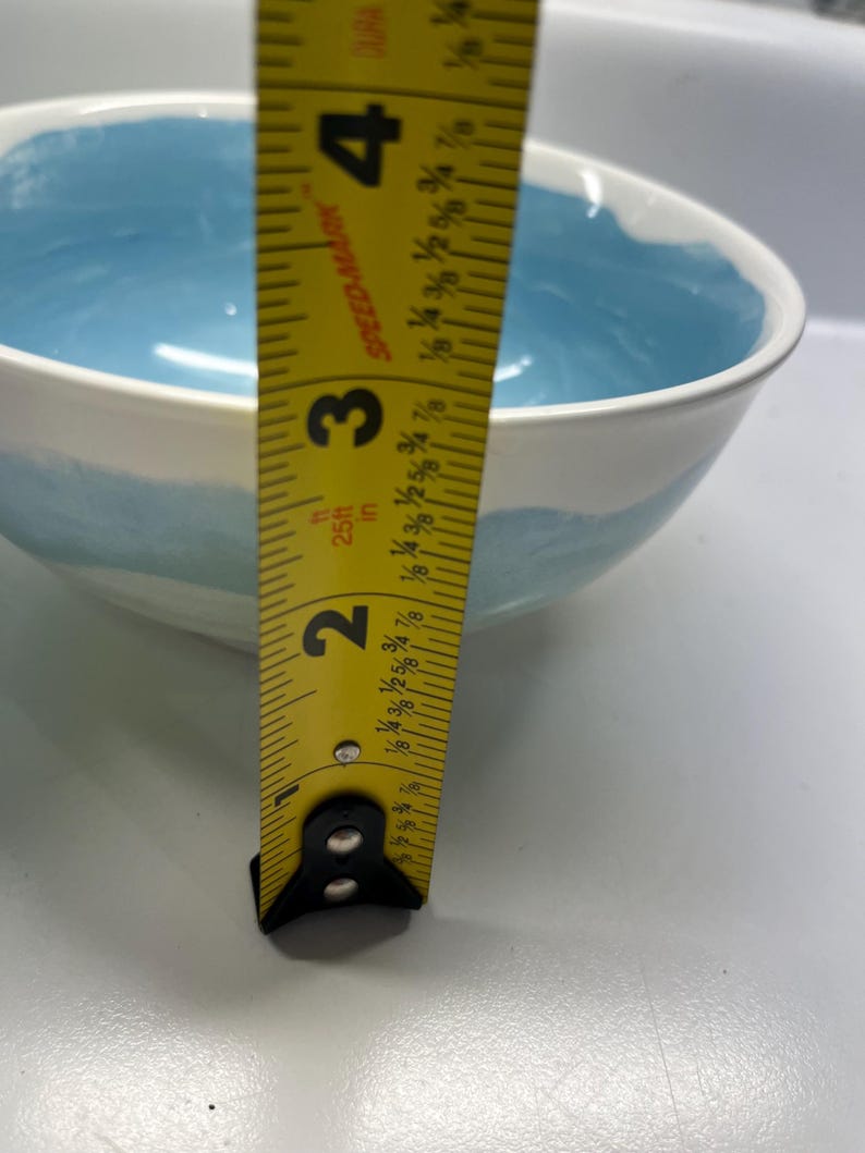 May include: A white ceramic bowl with a light blue interior and a blue painted band around the exterior. The bowl is approximately 7.6 cm deep, as measured by a yellow tape measure.