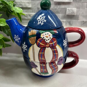 May include: A ceramic teapot and teacup set featuring a snowman design. The teapot is blue with white snowflakes and a green knob on the lid. The snowman has a red and blue plaid scarf and holds a broom. The teacup is red.