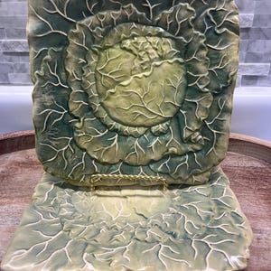 May include: A decorative ceramic cabbage-shaped plate and serving dish set. The plate is square with a raised cabbage design in shades of green and cream. The serving dish is rectangular with a matching cabbage leaf pattern.