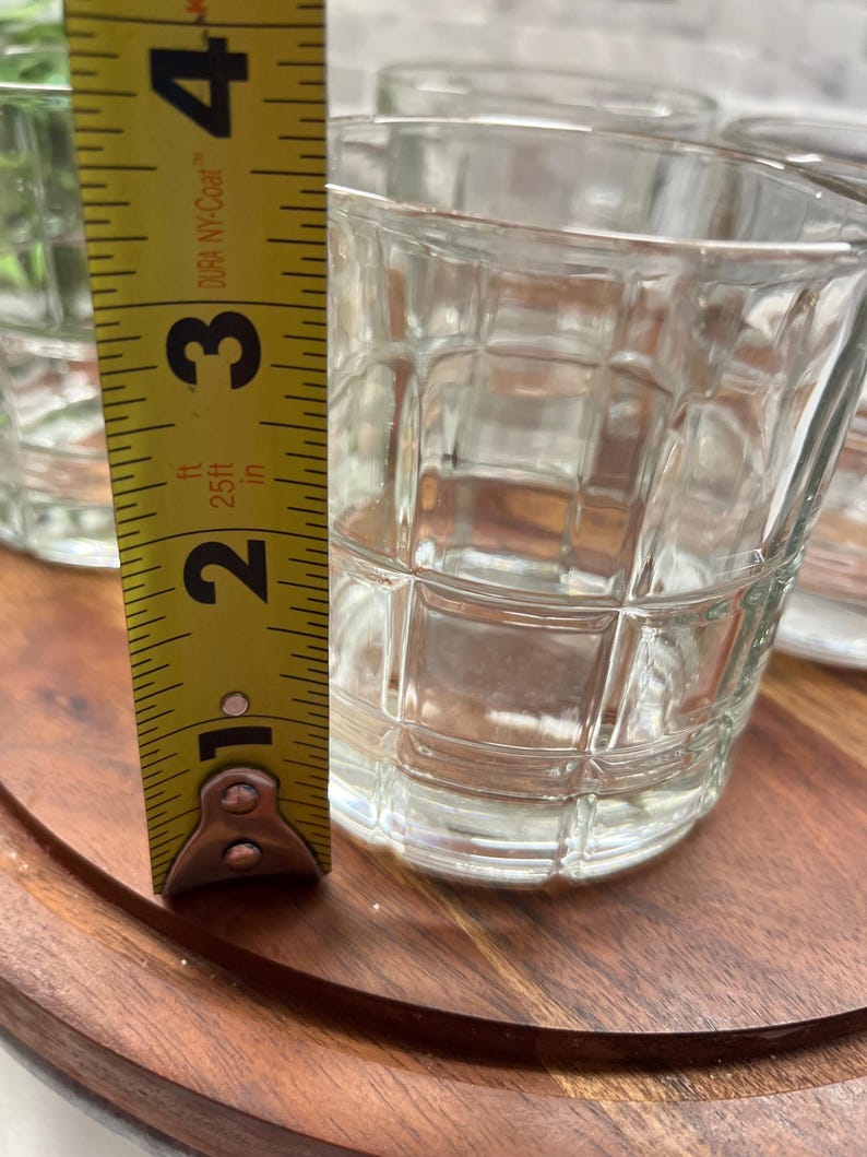Puede incluir: Conjunto de cuatro vasos transparentes de vidrio con un patr&oacute;n de cuadr&iacute;cula. Los vasos est&aacute;n sobre una superficie de madera. Se puede ver una cinta m&eacute;trica en la imagen, que muestra que la altura de los vasos es de 8,26 cm.