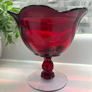 May include: A deep ruby red glass compote dish featuring a wide, scalloped rim and a rounded bowl. The vessel is supported by a decorative, multi-faceted pedestal stem that connects to a clear, circular glass base, creating a balanced silhouette.