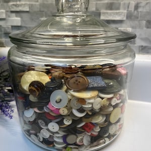 May include: A large, clear glass jar with a matching lid, filled with a diverse collection of assorted buttons in various shapes, sizes, and colors, including white, black, brown, blue, and cream, visible through the transparent glass walls.