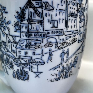Puede incluir: Taza de caf&eacute; de cer&aacute;mica blanca con una ilustraci&oacute;n azul y negra de una escena callejera parisina. El dise&ntilde;o incluye edificios, personas y el texto "IN ROUGE". La taza tiene un asa curva y un acabado liso y brillante.