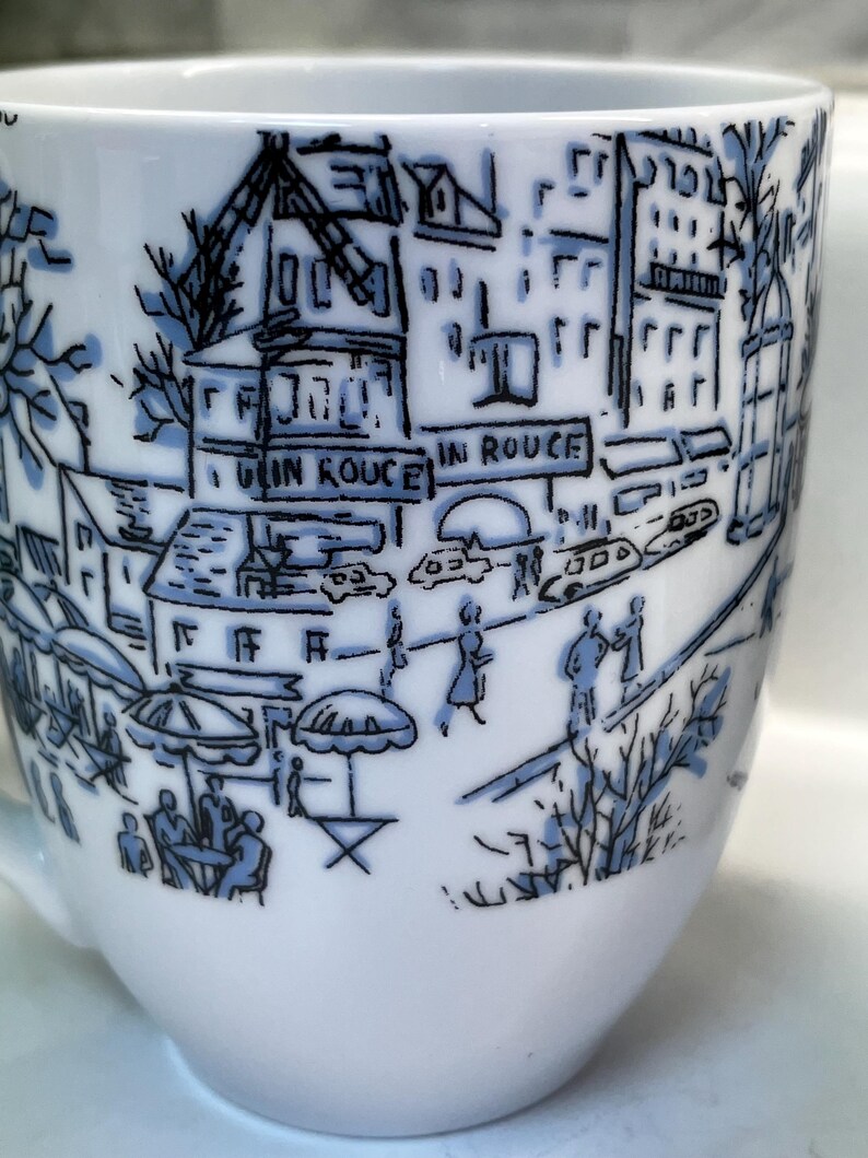 Puede incluir: Taza de caf&eacute; de cer&aacute;mica blanca con una ilustraci&oacute;n azul y negra de una escena callejera parisina. El dise&ntilde;o incluye edificios, personas y el texto "IN ROUGE". La taza tiene un asa curva y un acabado liso y brillante.