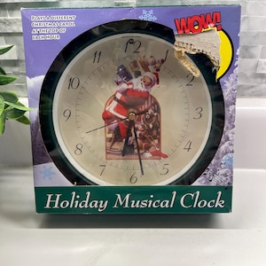 May include: A green-framed holiday musical clock with a festive Santa Claus design on the clock face. The clock plays a different Christmas carol at the top of each hour. The box reads "Holiday Musical Clock".