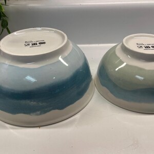 May include: Two ceramic bowls with a white base and a blue and teal watercolour design. The bowls are different sizes and have a glossy finish. The bowls are suitable for serving food.