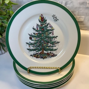 May include: A stack of white ceramic plates with a dark green rim. The center of the top plate features a detailed illustration of a decorated Christmas tree with a Santa Claus figure at the top and colorful wrapped gifts at the base.
