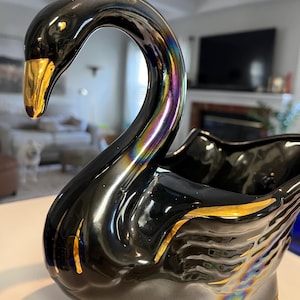 May include: A black ceramic swan-shaped planter with gold accents on the beak and wing details. The swan has a glossy finish with iridescent highlights. The planter is designed to hold plants or flowers.
