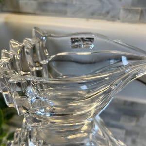 May include: A clear glass bowl with a sculptural, wave-like design and a jagged, stepped edge on one side. The glass is transparent and smooth, featuring a small, rectangular etched logo near the rim that appears to read as a brand mark.