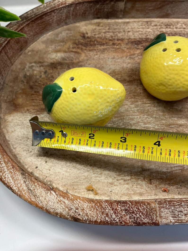 May include: A pair of ceramic lemon-shaped salt and pepper shakers. Each shaker is yellow with a green leaf and two or three holes on top. The shakers are on a wooden tray. The shakers are about 10 cm long.