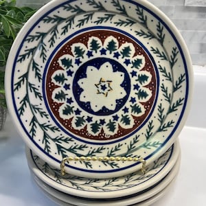 May include: A stack of four ceramic plates with a decorative design. The plates have a white base with a blue and green leaf border. The center features a brown, blue, and green pattern with a star and tree motif.