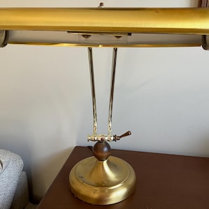 May include: A brass desk lamp featuring a wide, horizontal, tubular shade with a metallic gold finish. The lamp has a slender, dual-arm support structure connected to a circular base, accented by a dark brown wooden sphere at the central joint.