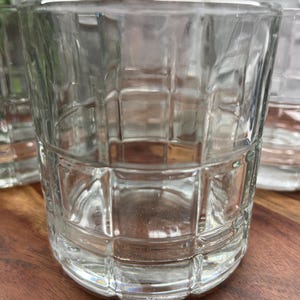 Puede incluir: Un juego de cuatro vasos transparentes de vidrio con un patr&oacute;n de cuadr&iacute;cula cuadrada. Los vasos est&aacute;n vac&iacute;os y se encuentran sobre una superficie de madera.