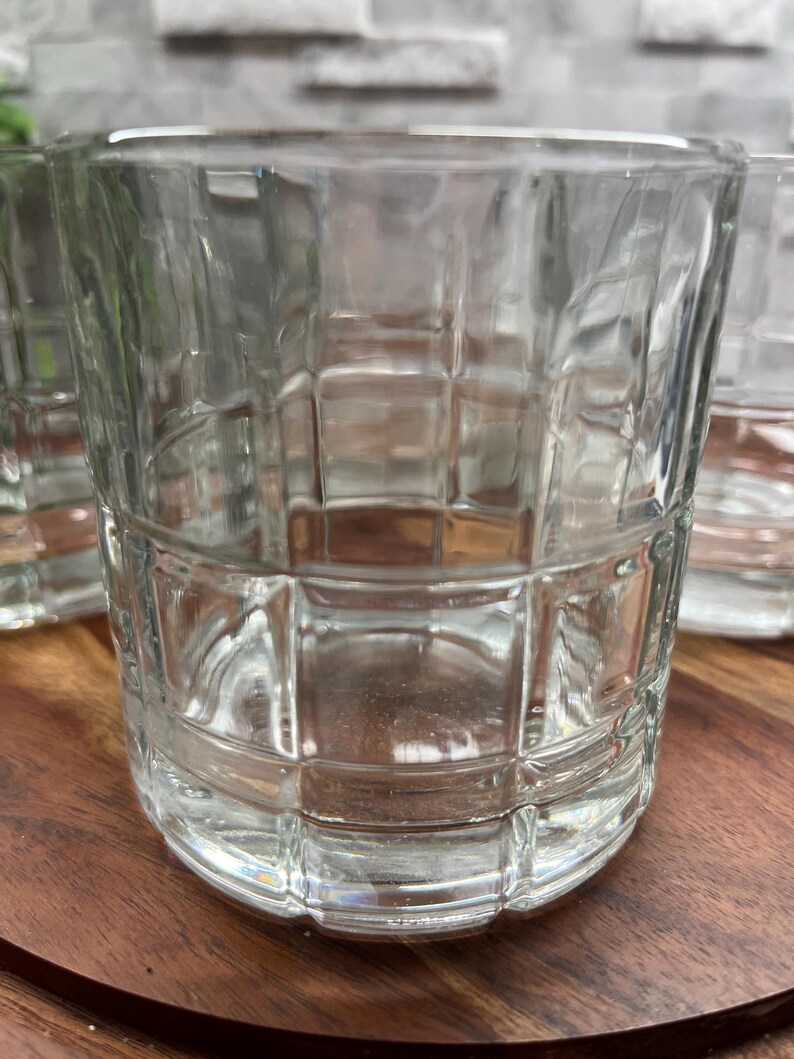 Puede incluir: Un juego de cuatro vasos transparentes de vidrio con un patr&oacute;n de cuadr&iacute;cula cuadrada. Los vasos est&aacute;n vac&iacute;os y se encuentran sobre una superficie de madera.