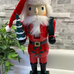 May include: A wooden Santa Claus nutcracker figurine. The figurine is painted red with black boots and a black belt. It holds a small green Christmas tree and wears a red hat with white trim. The base is green and gold.
