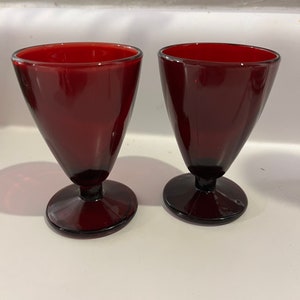 May include: Two ruby red glass goblets. Each goblet has a conical bowl, a short stem, and a circular base. The glasses are made of a dark red glass and are set against a white background.