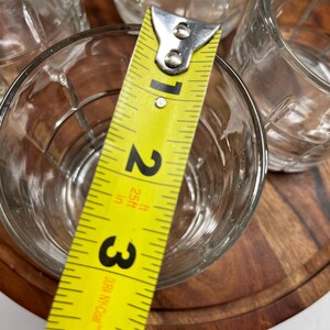 Puede incluir: Cuatro vasos transparentes de vidrio con un dise&ntilde;o texturizado, medidos con una cinta m&eacute;trica amarilla. La cinta m&eacute;trica indica 3 pulgadas.