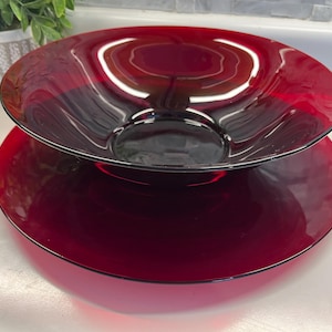 May include: A two-tiered, ruby-red glass serving dish. The top dish is a shallow bowl, and the bottom is a wide, flat plate. The glass has a glossy finish, reflecting light. The serving dish is likely for displaying or serving food.