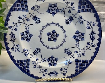 May include: A round ceramic plate featuring a white base with a dark blue floral pattern. The design includes a central star-shaped flower, surrounding blossoms, scrolling vines, and four corner sections with a blue grid pattern.