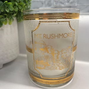 May include: A clear glass tumbler with a gold rim and gold detailing. The glass has an etched image of Mount Rushmore with the text "MT. RUSHMORE" on the front.