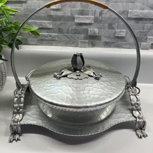 May include: A silver-colored serving dish with a hammered texture, a lid, and a handle with a woven grip. The dish sits on a matching tray, and the lid is topped with a floral design. The handle is arched and attached to the tray with decorative supports.