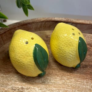 May include: Two ceramic lemon-shaped salt and pepper shakers. The shakers are yellow with green leaves and small holes on top. They are placed on a wooden tray.
