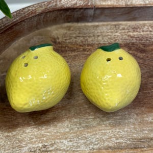 May include: Two yellow ceramic lemon-shaped salt and pepper shakers. Each shaker has a green stem and three holes. The shakers have a textured surface and are placed on a wooden tray.