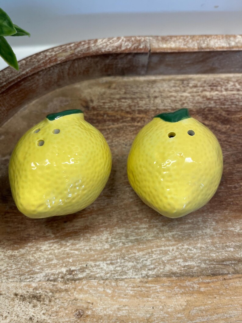 May include: Two yellow ceramic lemon-shaped salt and pepper shakers. Each shaker has a green stem and three holes. The shakers have a textured surface and are placed on a wooden tray.