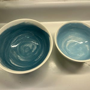 May include: Two ceramic bowls with a white exterior and a blue interior. The larger bowl has a dark blue, swirled design, while the smaller bowl has a lighter blue, swirled design. The bowls are likely for serving food.