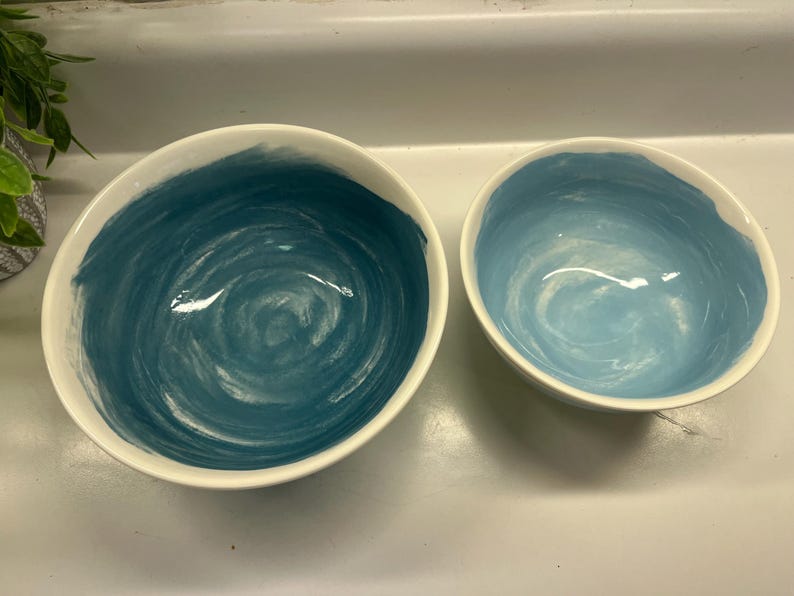 May include: Two ceramic bowls with a white exterior and a blue interior. The larger bowl has a dark blue, swirled design, while the smaller bowl has a lighter blue, swirled design. The bowls are likely for serving food.