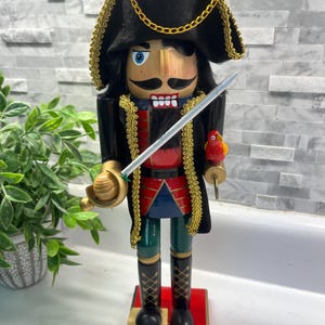 May include: A wooden nutcracker in the shape of a pirate, featuring a black hat with gold trim, a black coat, and a red and blue vest. The nutcracker holds a sword and a small parrot figurine.
