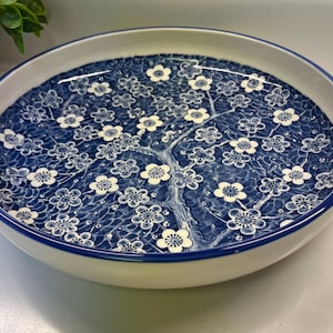 May include: A round, ceramic serving dish with a white rim and a blue floral design. The interior features a detailed pattern of white cherry blossoms on a blue background, creating a decorative and functional piece for serving food.