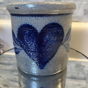 May include: A cylindrical stoneware crock featuring a textured gray speckled finish and a prominent cobalt blue hand-painted heart design on the front. The vessel has a thick, rounded rim and a straight-sided body with a rustic, porous appearance.