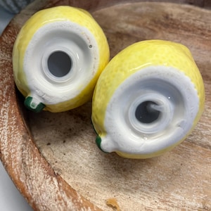 May include: Two ceramic lemon-shaped salt and pepper shakers. The shakers are yellow with white bases and green stems. They are sitting on a wooden tray.
