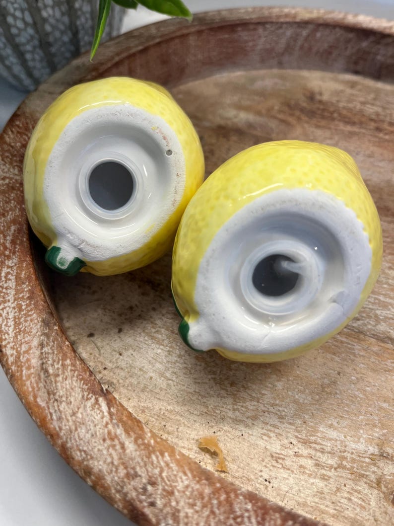 May include: Two ceramic lemon-shaped salt and pepper shakers. The shakers are yellow with white bases and green stems. They are sitting on a wooden tray.