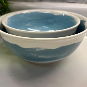 May include: Two ceramic bowls, one inside the other. The bowls are white with a band of blue paint around the exterior and interior rims. These bowls are suitable for serving or mixing.