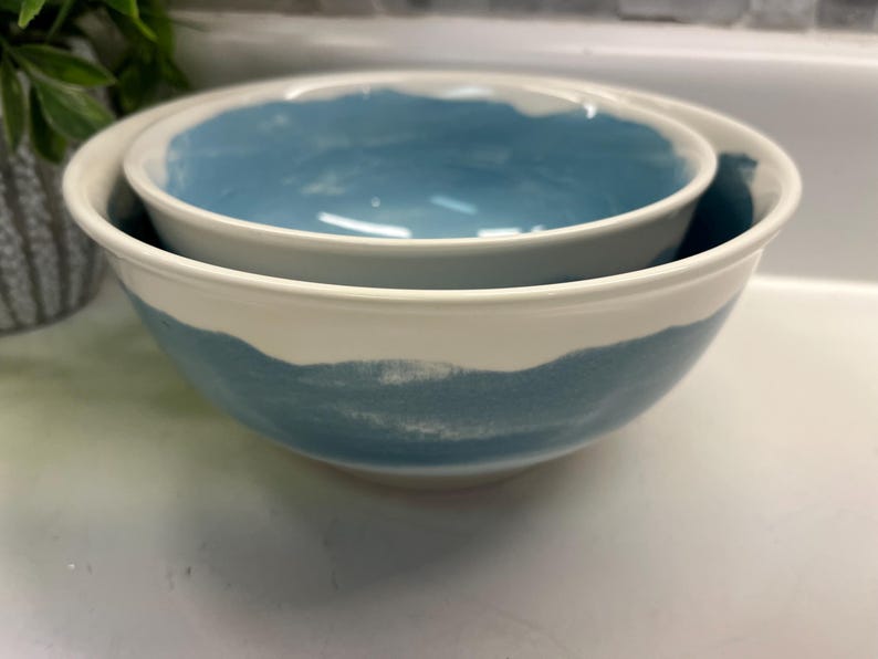 May include: Two ceramic bowls, one inside the other. The bowls are white with a band of blue paint around the exterior and interior rims. These bowls are suitable for serving or mixing.