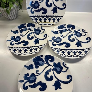 May include: Set of four white ceramic plates with a blue floral design. The plates feature a repeating pattern of stylized flowers and vines, with a decorative border. The plates are arranged on a white surface.