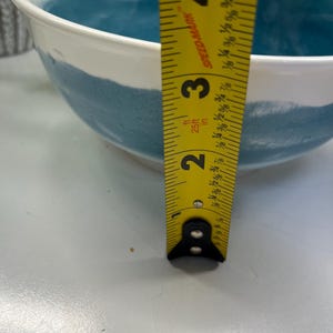 May include: White ceramic bowl with a teal blue painted band. A yellow measuring tape shows the bowl is approximately 10 cm deep. The bowl has a smooth, glossy finish and is suitable for serving food.